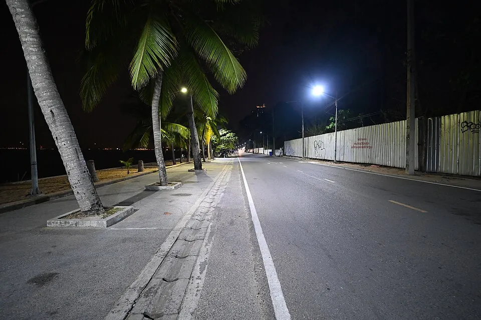 A deserted coastal road stretches into the night in Klet Kaeo, Sattahip District, illuminated by the cool white glow of overhead street lamp