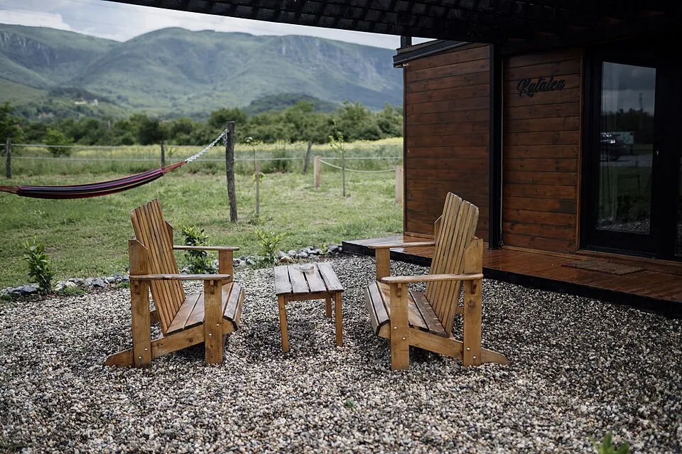 Two wooden Adirondack chairs and a small table sit on gravel, facing picturesque mountains under a cloudy sky. Green fields and trees surrou